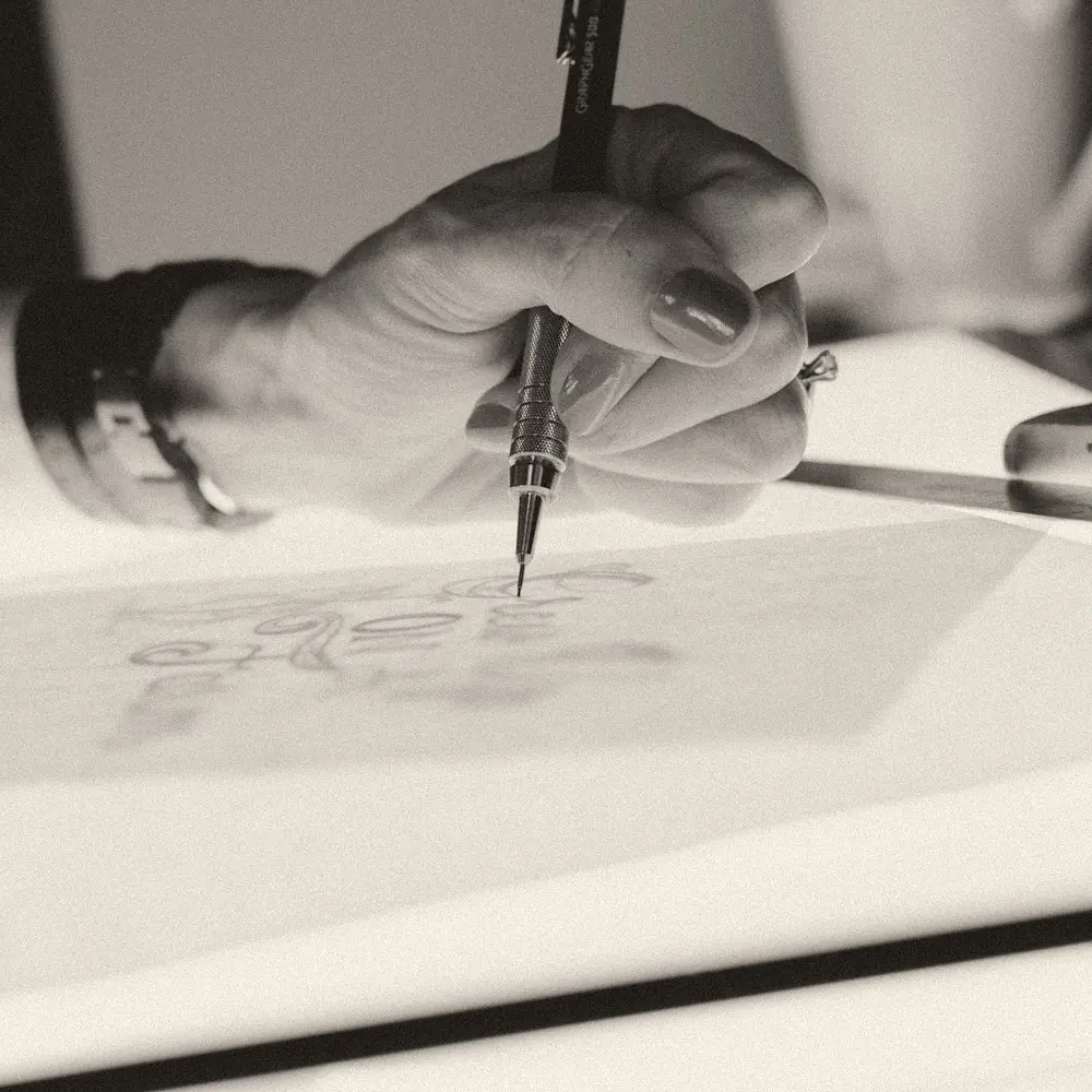 A hand tracing a sketch with a fine pen on tracing paper over a light table